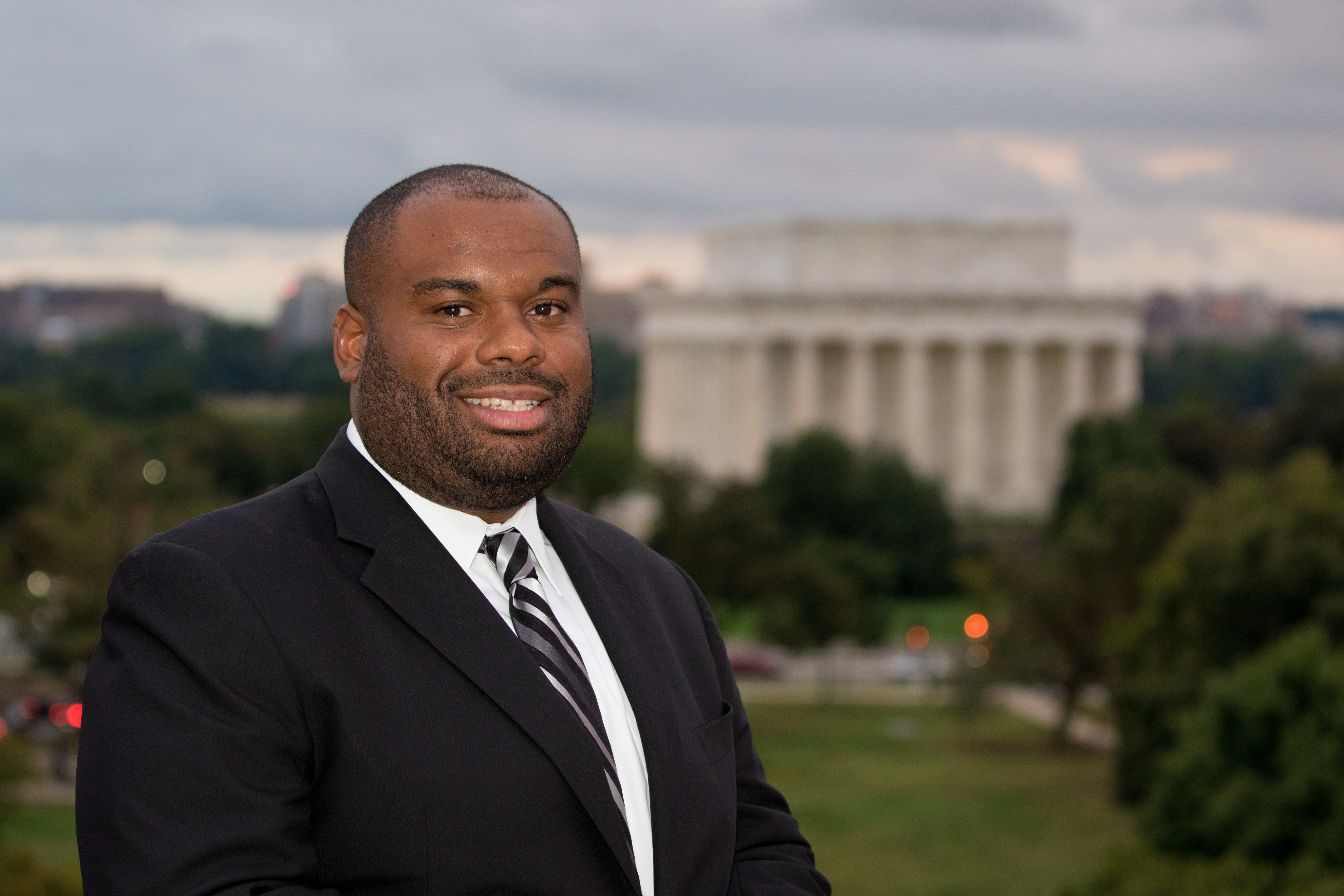 A man smiling in a dark suit, white shirt, and dark striped tie, with the Lincoln Memorial in the distance